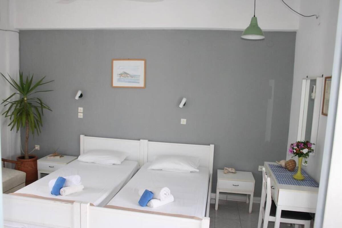 A hotel room with two beds, a desk, a chair, a small table with a vase, and a potted plant.