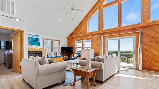 A spacious living room with a high ceiling, wooden walls, and large windows offering a view of the beach. - To the Beach and Back at Salvo 5 Bedroom Home with a Private Pool, Spa and Near Beach Access! (Salvo)