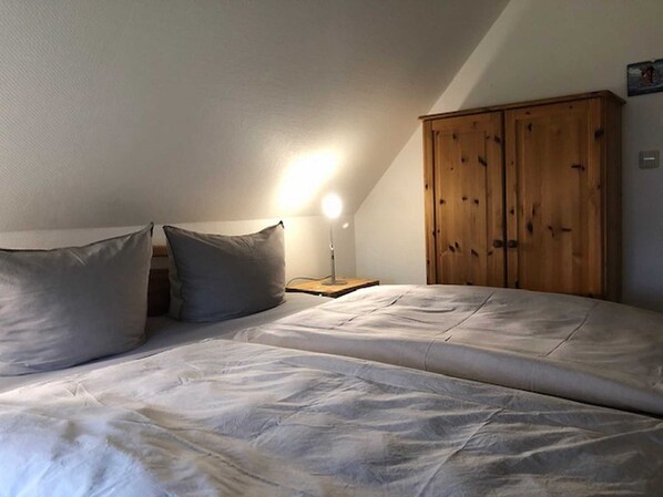 A bedroom with a bed, two pillows, a nightstand, a lamp, and a wooden wardrobe. - Apartments Gueren - Kasparhus (Insel Hiddensee)