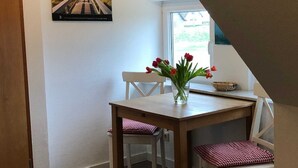 A dining area with a wooden table and chairs, a vase with red flowers, and a window with a view of greenery.
