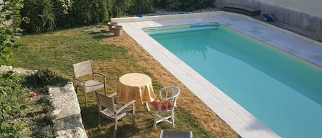 Una piscina rectangular con solárium alicatado, rodeada de césped, un muro de piedra, una mesa redonda, dos sillas y una tumbona.