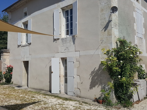Ein zweistöckiges Haus mit weißen Fensterläden, ein kleiner Garten mit Topfpflanzen und eine sichtbare Tür.