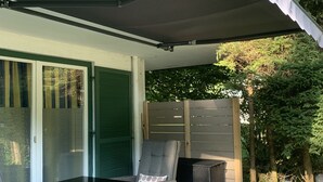 Eine überdachte Terrasse mit Korbmöbeln und einem Tisch, ein Holzzaun und eine Statue im Hintergrund.