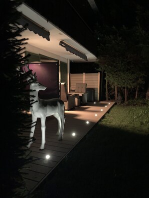 Un sendero conduce a un espacio al aire libre techado con una silla y una mesa, rodeado de árboles y una escultura de un ciervo.