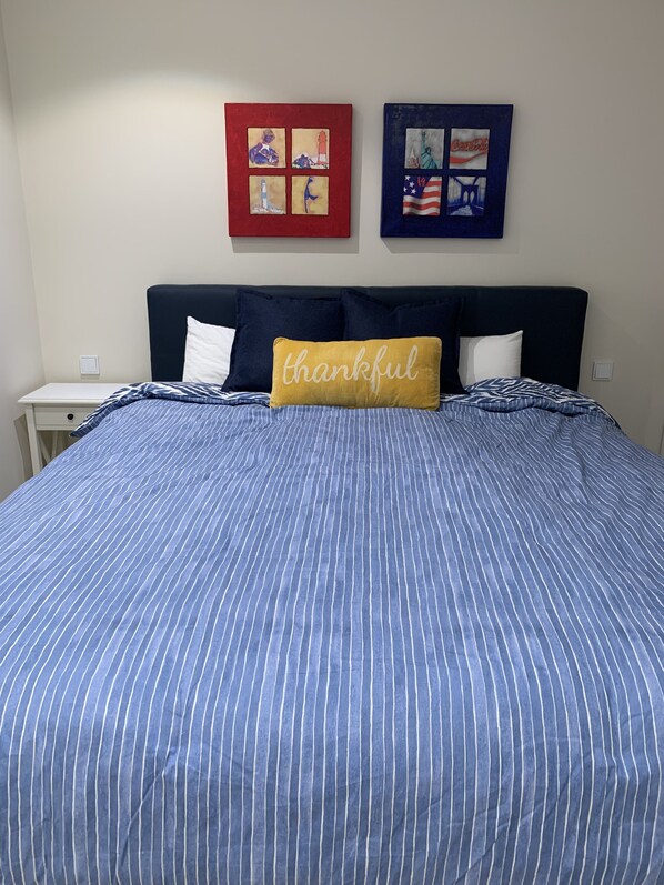 Una cama bien hecha con un edredón azul y blanco a rayas, una almohada amarilla con la palabra 'thankful' y dos obras de arte en la pared que representan monumentos emblemáticos de Estados Unidos.