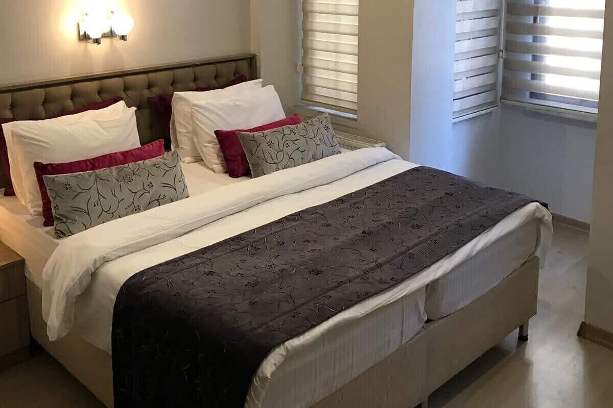 Une chambre à coucher comprenant un lit, des tables de chevet, un luminaire fixé au mur et une fenêtre avec des stores.