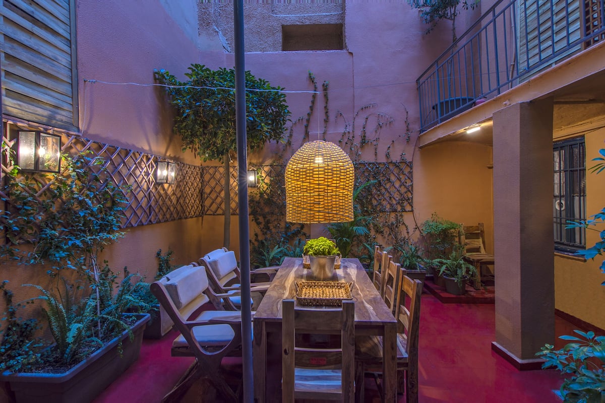 An outdoor dining area with a wooden table and chairs, surrounded by potted plants and a decorative lamp.
