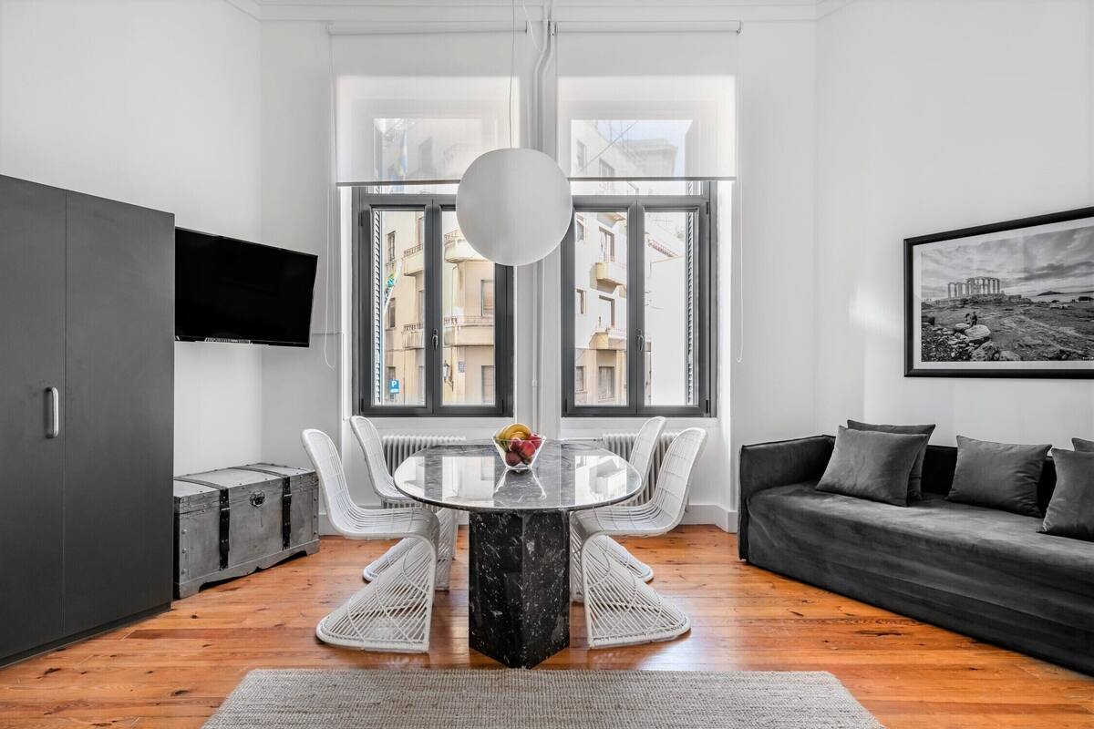 A modern living room with a round glass table, white wicker chairs, a dark sofa, and a mounted TV.