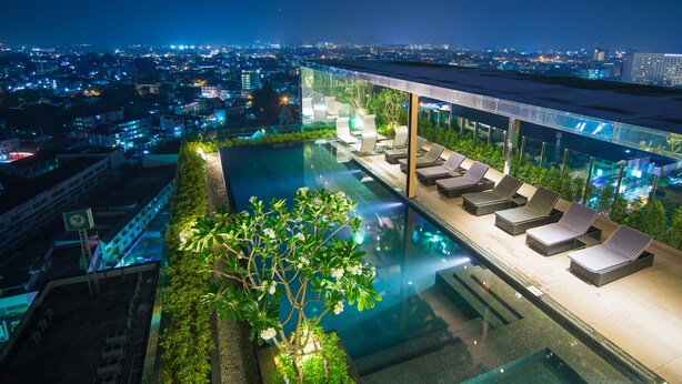 A rooftop infinity pool with sun loungers, surrounded by greenery and overlooking a cityscape at night. - The Astra Condo (Chiang Mai)