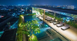 A rooftop infinity pool with sun loungers, surrounded by greenery and overlooking a cityscape at night. - The Astra Condo (Chiang Mai)