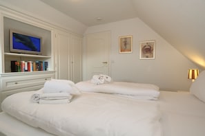 Une chambre d’hôtel avec un téléviseur posé sur une étagère, un lit avec du linge de maison blanc, une porte et deux tableaux encadrés au mur.