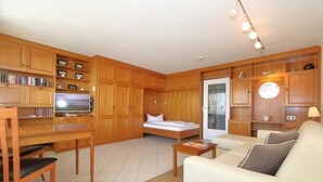 A compact living space with a bed, sofa, wooden cabinets, a television, and a dining area. - Haus am Meer14-app 091 WB (Sylt)