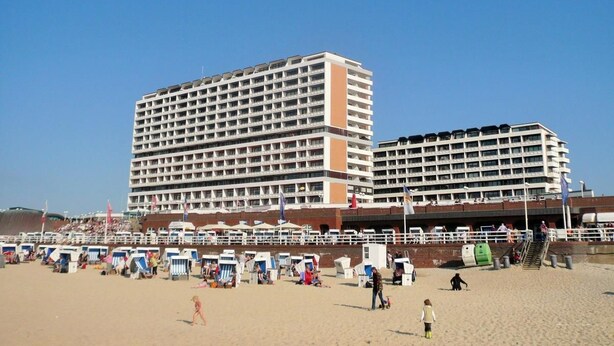 A large hotel with multiple floors and balconies, a sandy beach in front, and beach chairs and umbrellas. - Haus am Meer14-app 091 WB (Sylt)