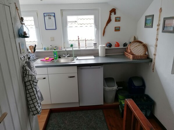Una cocina con un lavamanos, un escurreplatos, un cubo de basura y una ventana.