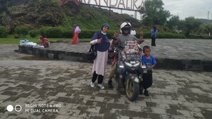 A sign with 'Kuta Mandalika' written on it, a paved area, and a motorcycle with a child sitting on it. - T&T Homestay (Kuta)