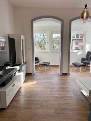 Un salon moderne avec un téléviseur à écran plat, un canapé et deux fauteuils. Il y a deux grandes fenêtres donnant sur des arbres et des immeubles.