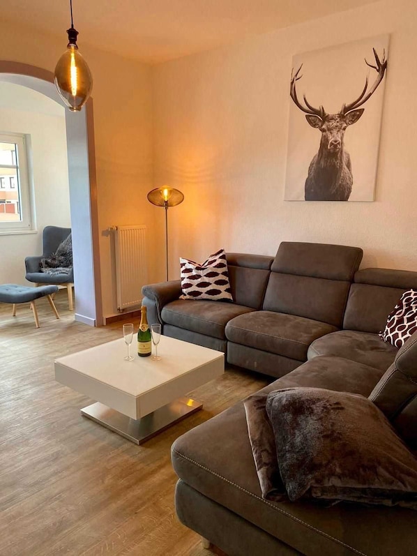 Un salon moderne avec un canapé d’angle, une table basse sur laquelle se trouvent une bouteille et un verre, une lampe sur pied et une œuvre d’art représentant une tête de cerf accrochée au mur.