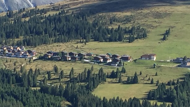 A mountainous village with houses clustered on a hillside, surrounded by dense forests and a backdrop of mountains.