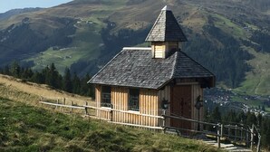 A wooden cabin with a steep roof, surrounded by a wooden fence, set against a mountainous backdrop.