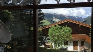 A wooden cabin with a sloped roof, surrounded by greenery and mountains in the background.