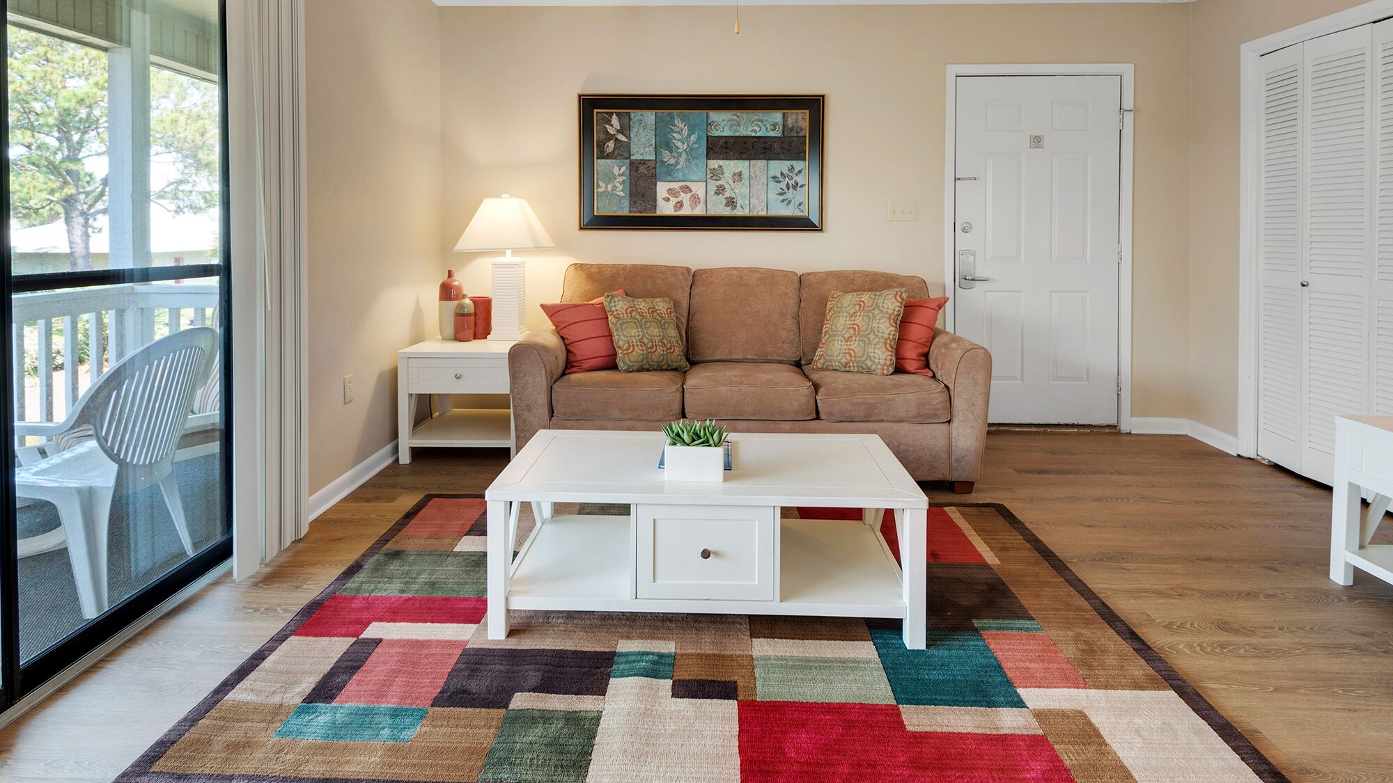 A living room with a beige sofa, a white coffee table, and a colorful rug. There is a framed artwork on the wall and a door to the right.