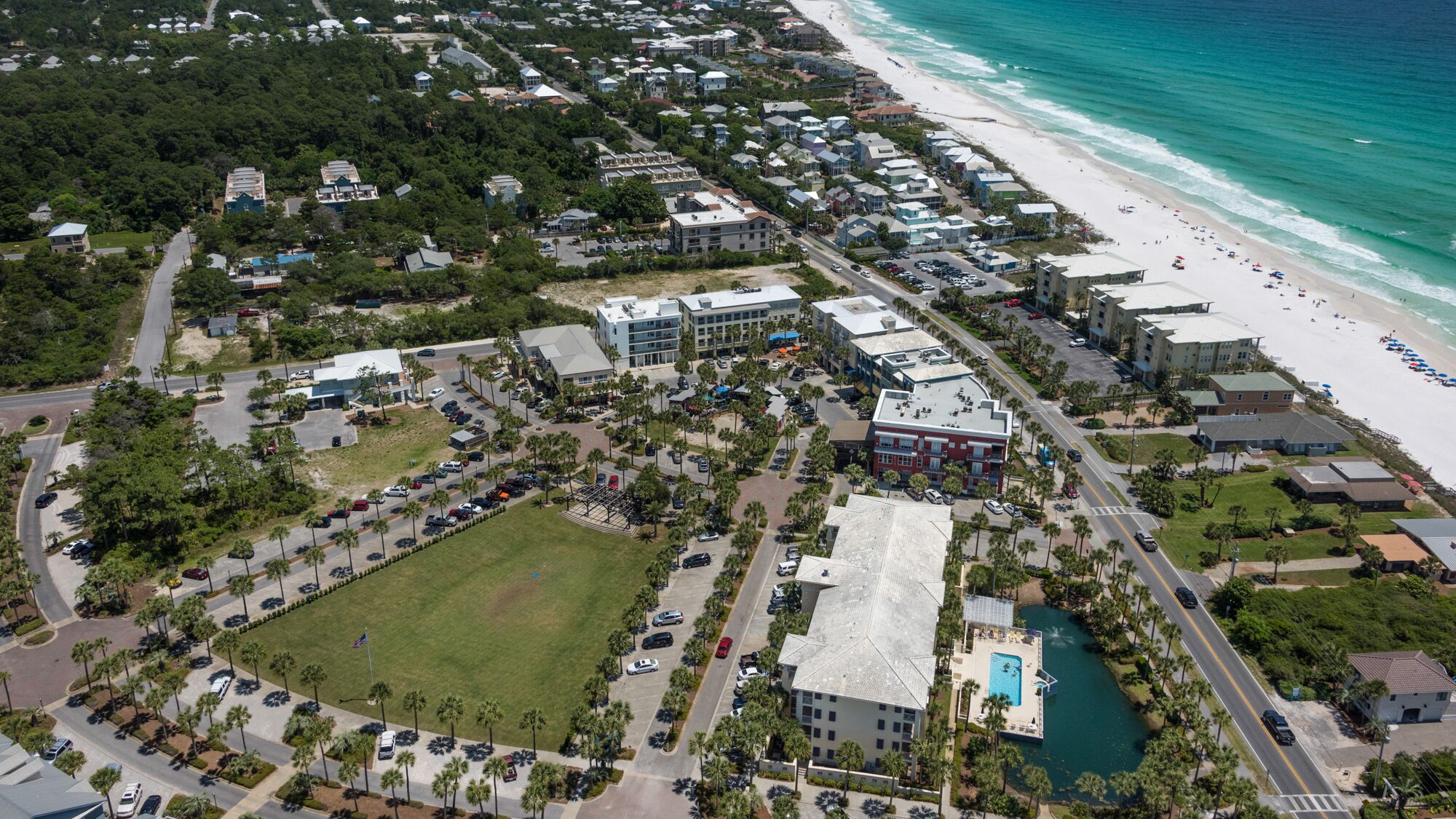 Luftaufnahme eines Küstenresorts mit großem Pool, Strandhäusern und einem zentralen Platz mit Parkplätzen.