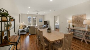 A spacious living area with a wooden dining table, comfortable seating, a ceiling fan, and a view through a large window.