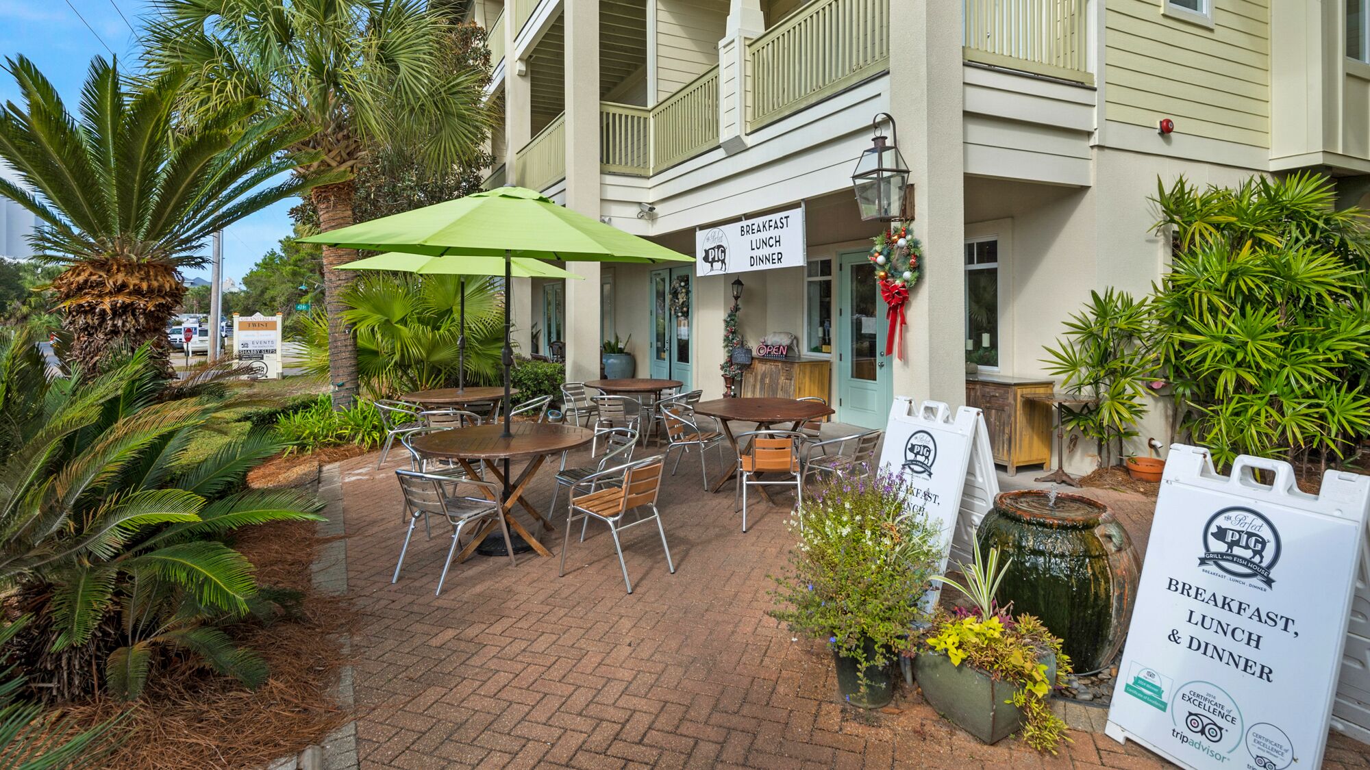 Un patio exterior con mesas y sillas, un paraguas verde y un letrero de restaurante que ofrece desayuno, comida y cena.