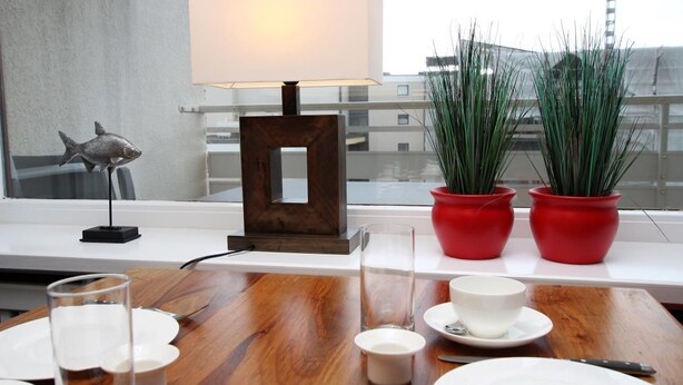 A dining table set with plates, glasses, and cutlery, a lamp, and two red potted plants. - Hussmann, Germany (Sylt)