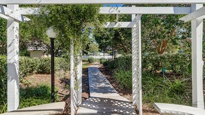 Een tuinpad onder een witte pergola met een rasterontwerp, omringd door groen en een heldere lucht.