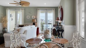 Ein Wohnzimmer mit einem weißen Sofa, einem Glastisch und Korbstühlen. Es gibt einen Deckenventilator, ein Fenster mit Jalousien und einen Blick auf Häuser im Freien.