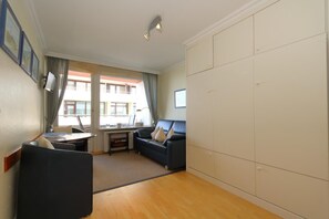 Ett litet rum med en soffa, ett litet bord och ett skrivbord med en stol. Det finns en stor garderob och ett fönster med gardiner.