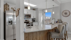 Eine moderne Küche mit einem Edelstahlkühlschrank, einer Mikrowelle und einer Kaffeemaschine. Eine Holzeinsel mit Barhocker und eine große Uhr an der Wand sind vorhanden.