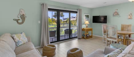 A spacious living room with a large sliding glass door leading to a balcony, a flat-screen TV mounted on the wall, and a small wooden table with a lamp.