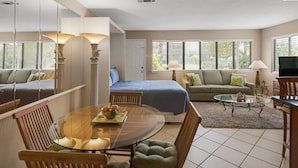 A well-lit, spacious room with a dining area, a sofa set, a television, and large windows with blinds.