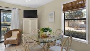 Un coin repas avec une table en verre entourée de chaises en osier, d’un canapé en osier et d’un téléviseur fixé au mur.