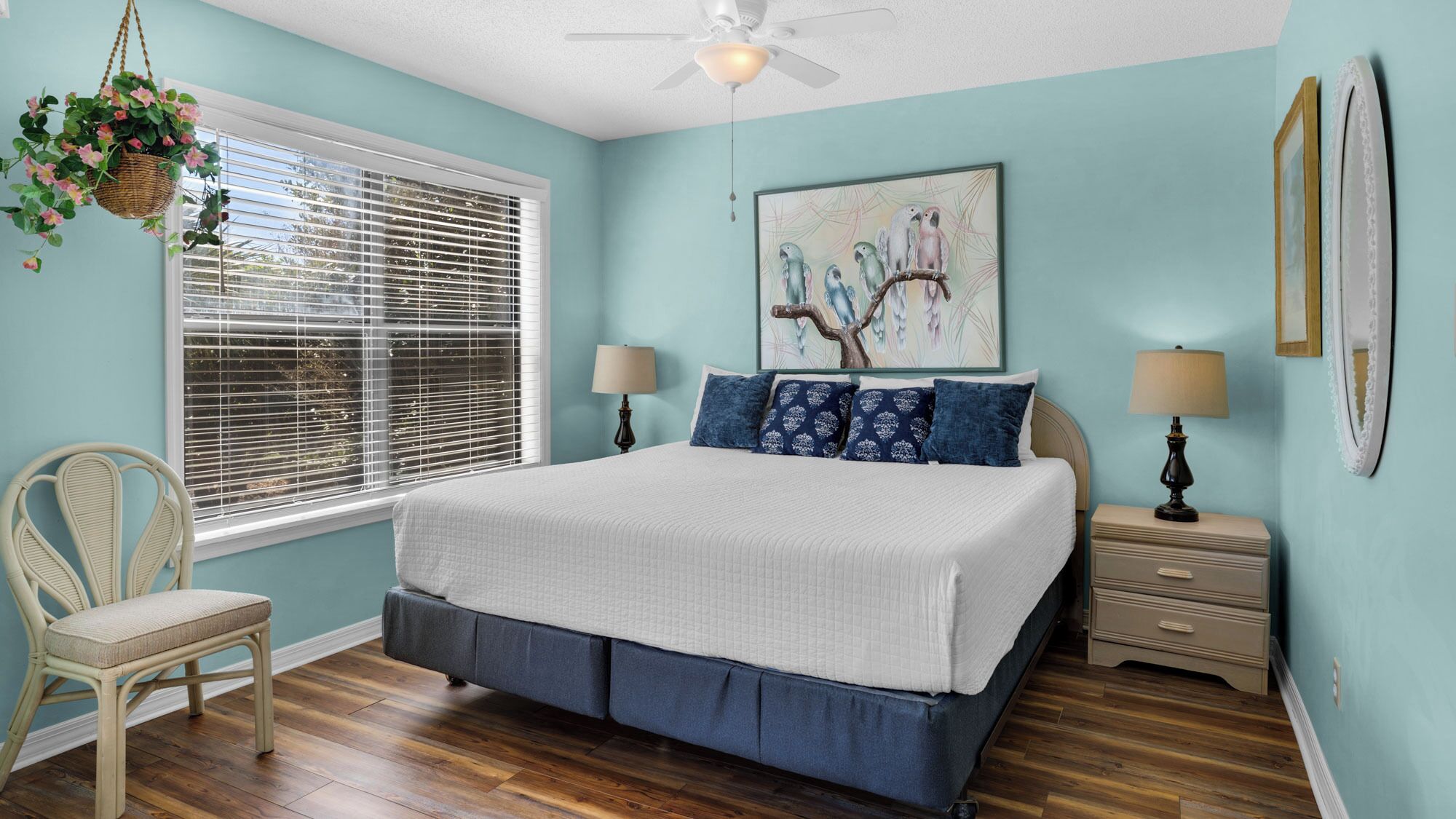 A bedroom with a bed, two bedside tables, a chair, a hanging plant, and a window with blinds.