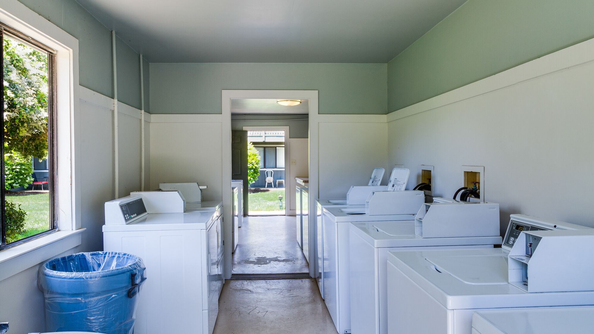A laundry room with multiple washing machines, a blue trash bin, and a view of a backyard through a window.