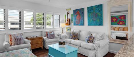A living room with a light grey sofa, a blue coffee table, and a wooden chest of drawers. There are two paintings on the wall, a guitar, and a lamp on a side table.