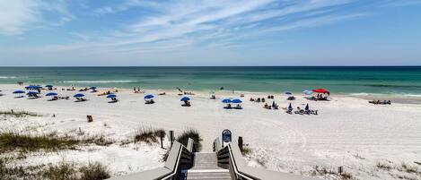 A beach with a wooden staircase leading down to the sand, numerous beach umbrellas, and a clear view of the ocean.