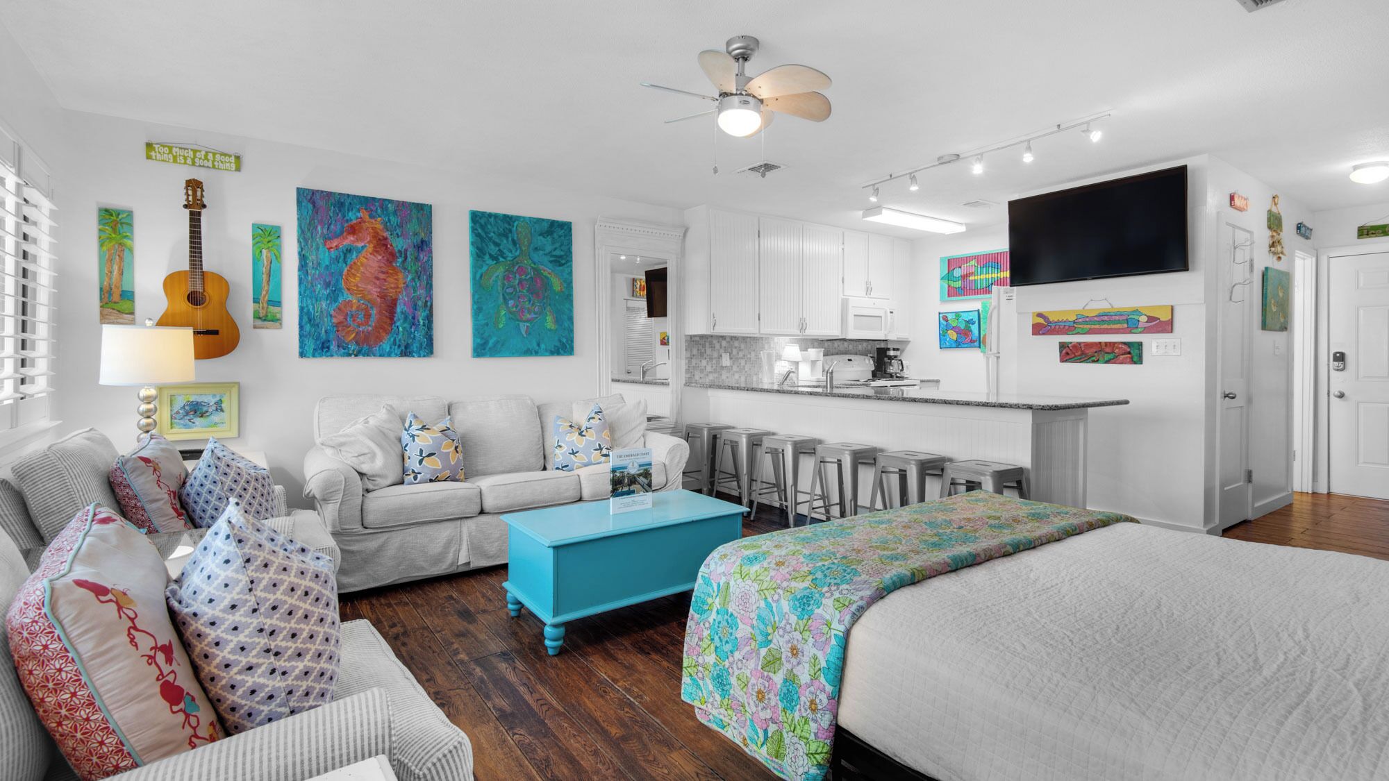 Una sala de estar moderna y bien iluminada, con sofá, mesa de centro y cama. Se observa una zona de cocina con mostrador y taburetes.