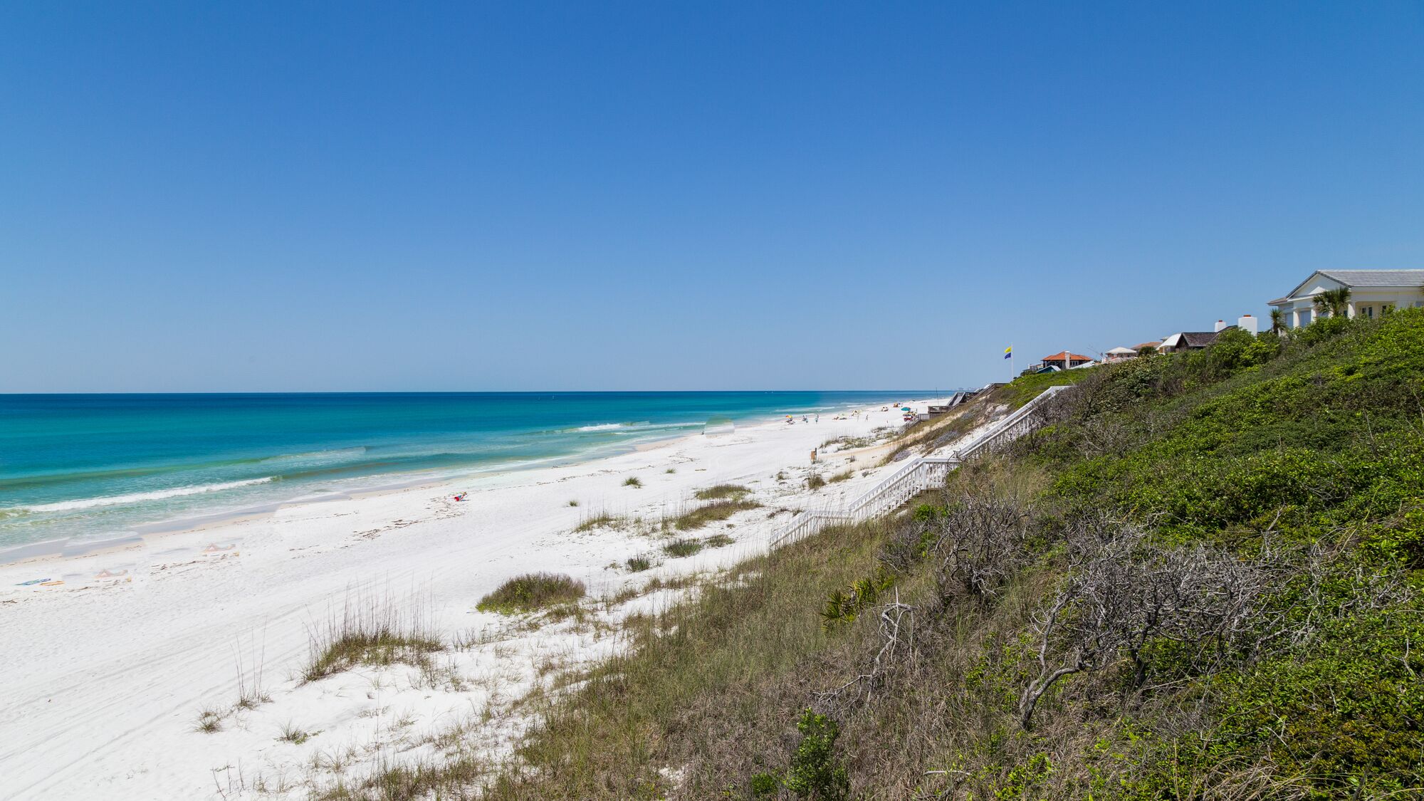 Une plage avec un rivage de sable blanc, une eau d’un bleu limpide et une maison située sur une colline, d’où l’on a une vue sur la plage.
