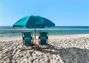 Dos sillas de playa con la marca 'Beachcomber' en una playa arenosa con un paraguas azul grande.