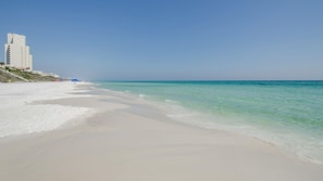 Ein Strand mit klarem türkisblauem Wasser, einem sandigen Ufer und einem hohen, weißen Gebäude im Hintergrund.