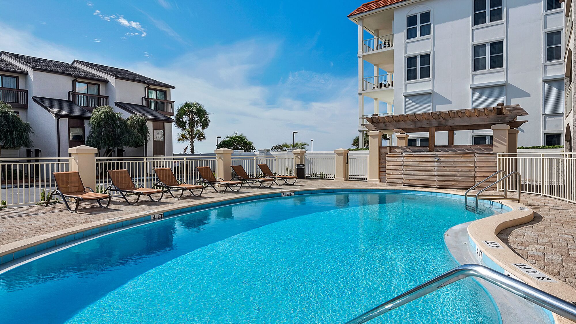 A swimming pool with lounge chairs and a pergola, surrounded by a fence and buildings.