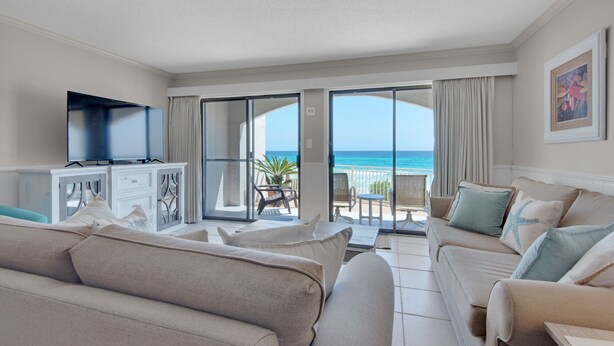 Ein geräumiges Wohnzimmer mit einem großen Flachbildfernseher, beigen Sofas und einem Balkon mit Meerblick.