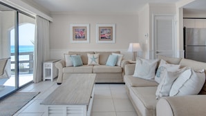 Ein Wohnzimmer mit einem beigen Sofa, einem Holztisch und Blick auf das Meer durch eine Schiebetür. An der Wand hängen zwei gerahmte Kunstwerke, und auf einem Beistelltisch steht eine Lampe.