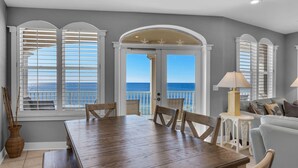 A dining area with a wooden table and chairs, a sofa, and a lamp. There are large windows with shutters and a view of the ocean.
