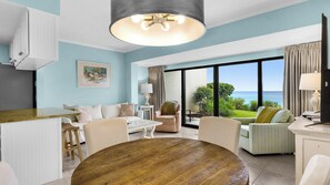 A modern living room with a wooden dining table, a sofa, and a chair. There is a large window with a view of the ocean. - Gulf Front with Blue Views, Pool, Great Location & Free Attraction Tickets! (Santa Rosa Beach)