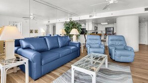 Una sala de estar moderna con un sofá azul, dos sillones, una mesa de centro de vidrio y una planta grande en maceta.